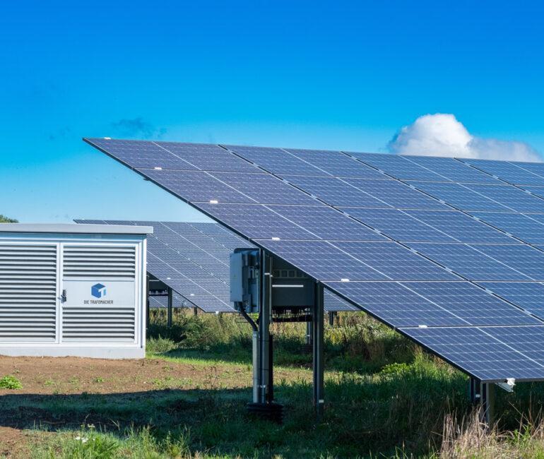 Leicht vergilbte Wiese, auf der ein Solarpark mit Wechselstation installiert wurde. Es ist strahlend schönes Wetter.