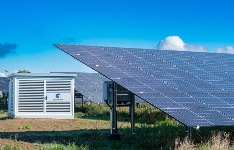 Leicht vergilbte Wiese, auf der ein Solarpark mit Wechselstation installiert wurde. Es ist strahlend schönes Wetter.